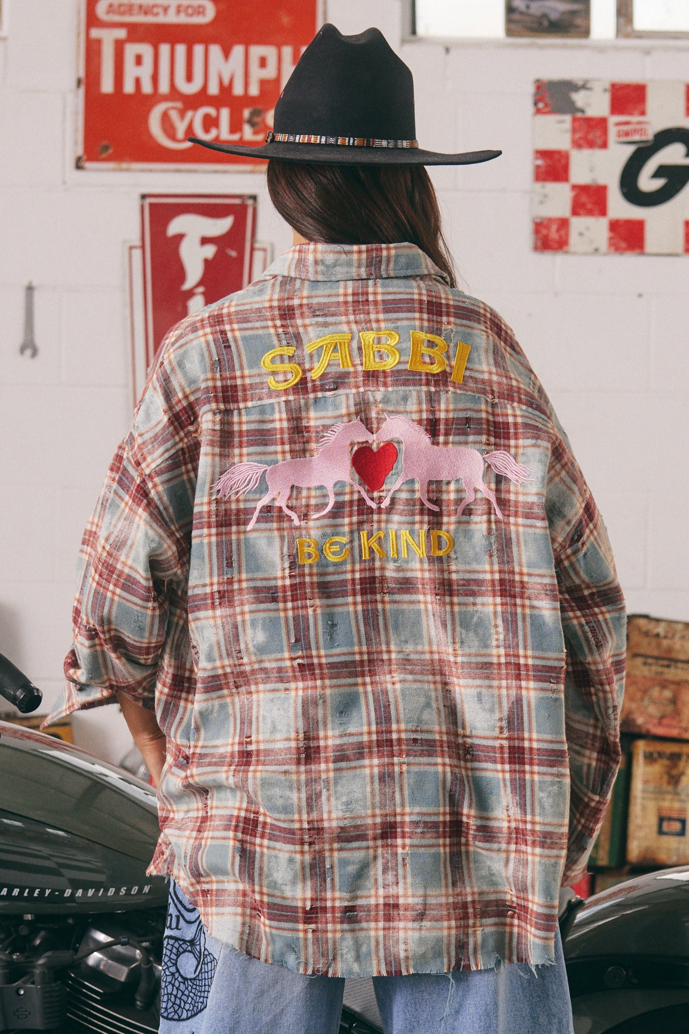 The Horse Power Distressed Flannel Shirt – Burgundy
Where grit meets kindness. The Horse Power Distressed Flannel in Burgundy is your go-to layer for laid-back rebellion. With a sun-faded, vintage-inspired burgundy and blue check, this oversized shirt brings serious attitude with a soft side.
The back steals the show with SABBI’s signature embroidered pink ponies, chenille red heart, and “BE KIND” slogan — a sweet reminder stitched into a tough-as-nails piece. The right sleeve is embroidered with “HORSE P