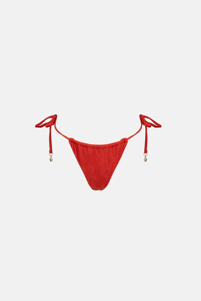 The Leilani Gathered Tie Side Its Pant in Red offers minimal coverage with functional gathers at the front and back for a customizable fit. Metal ring detail at the side string ties adds a unique flair, while adjustable string ties with beads on the ends and zig-zag stitching at the leg openings elevate its design. The back pant panel is lined in self-fabric for extra comfort, complemented by the metal coin logo.
