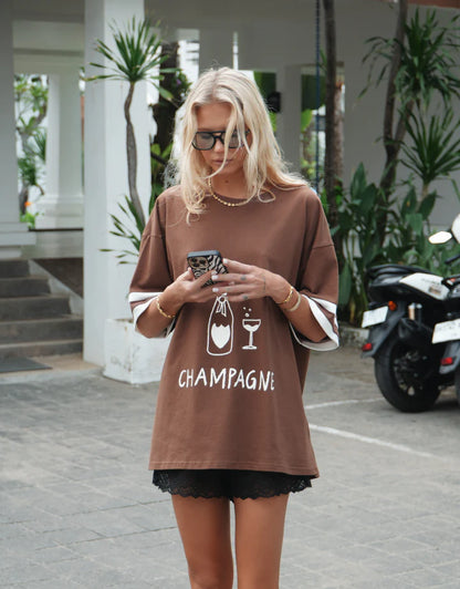 Casual comfort meets effortless style with The Champagne Tee — a versatile wardrobe essential designed for everyday wear.

Crafted on a warm brown base, this tee features subtle ribbing on the sleeves for a touch of texture and shape, while the bold front print adds personality and edge. The relaxed fit makes it easy to style with denim, skirts, or shorts, giving you an elevated casual look that works day or night.