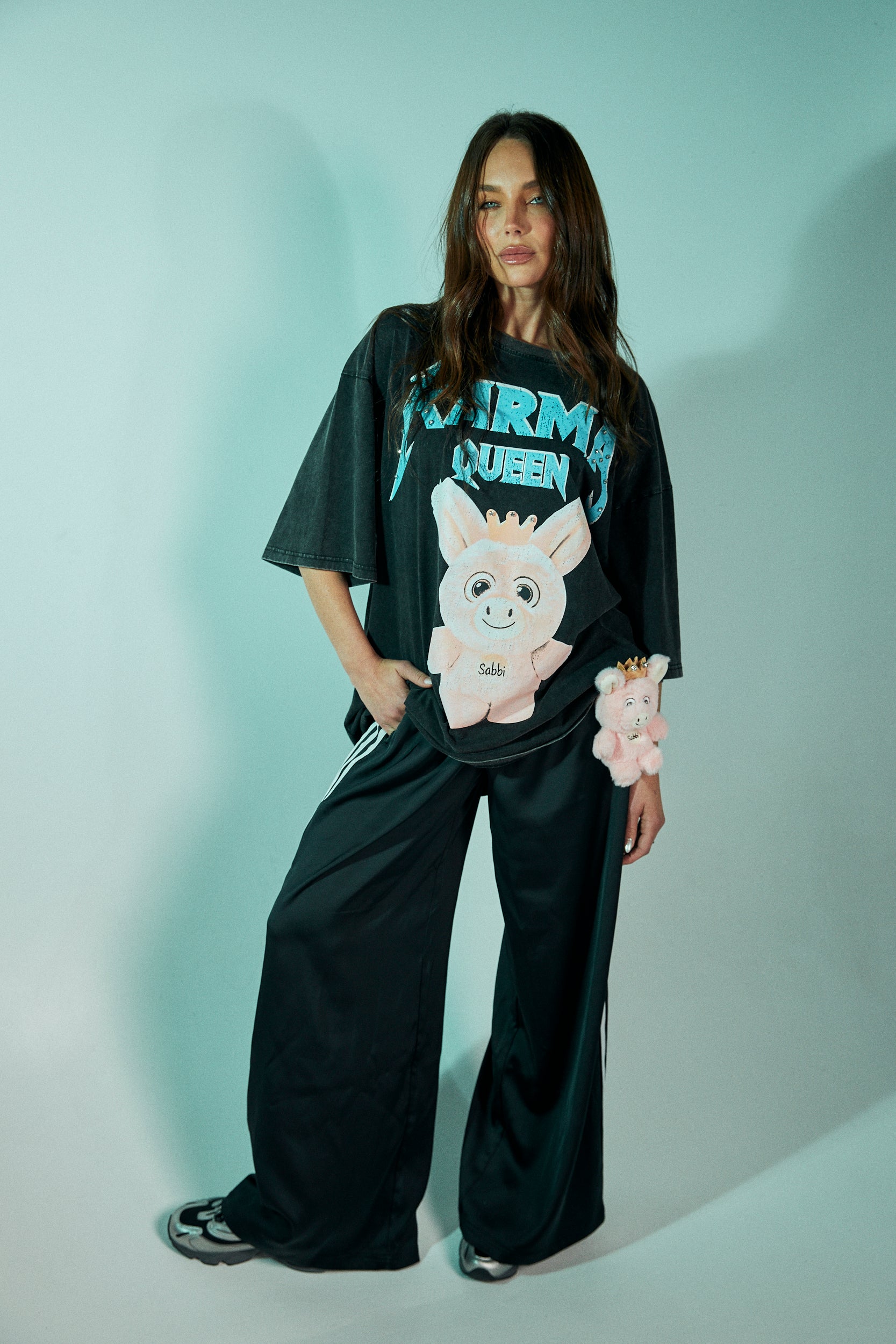 Rule your world in the Karma Queen Bling Tee – where sass meets sparkle. Crafted from premium black cotton with a vintage wash, this oversized tee features a bold graphic of the Karma Queen piglet, crowned in rhinestones for the perfect touch of bling. The statement “KARMA QUEEN” shimmers across the chest in studded detail, while the back reads “I SAW THAT – KARMA” as a cheeky reminder that nothing goes unnoticed.
