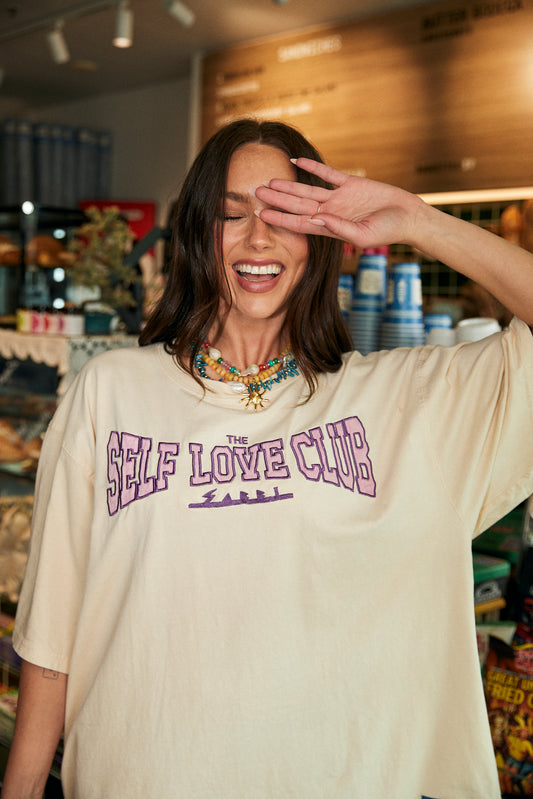 Step into unapologetic comfort and bold self-expression with The Very Oversized Self Love Club Tee in Off White. Crafted from premium, breathable cotton, this tee is designed with an ultra-relaxed, slouchy fit that drapes effortlessly – perfect for layering or rocking solo.

The front features the iconic SELF LOVE CLUB graphic in striking purple, a wearable reminder to put yourself first. On the back, a powerful pink statement – “The right people love the parts you try and hide” – serves as both a pep talk 