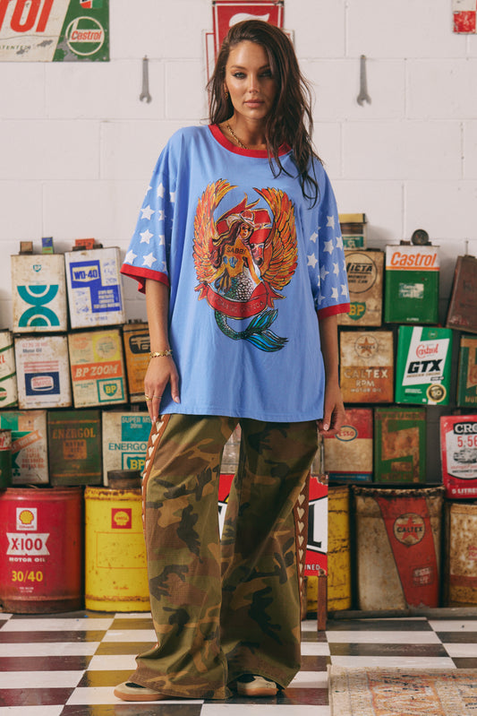 Born to stand out. The Dream Big Brenda Tee in Periwinkle is a fearless blend of retro biker energy and bold feminine power. Featuring the iconic winged mermaid front print and bold varsity-inspired back graphic, this oversized tee is made for wild hearts who ride loud and dream louder.

Designed in a vibrant periwinkle blue with contrast red ribbed trims and all-over star-print sleeves, it’s a punch of colour, confidence, and comfort in one seriously cool silhouette.