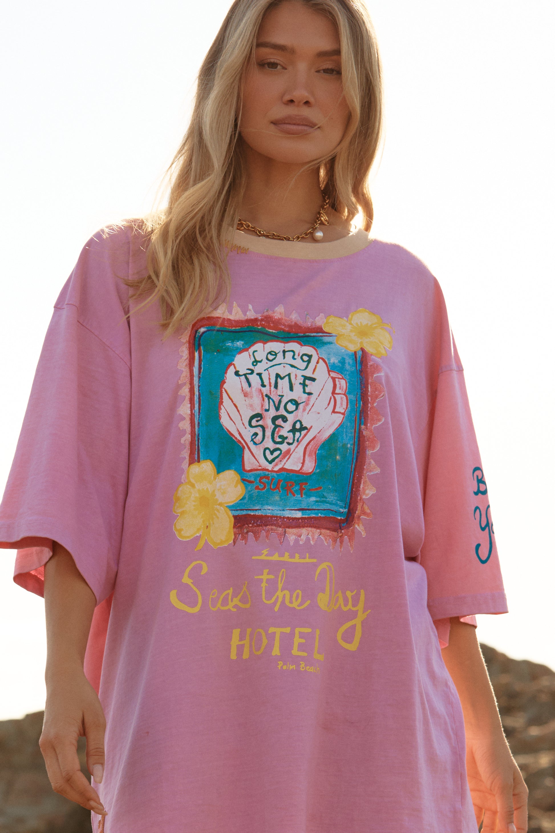 Sabbi - Meet your new off-duty essential. The Seas The Day Hotel Tee – Pink is all about laid-back beach energy with a playful, feel-good twist. Washed in a sun-faded pink and finished with hand-drawn coastal artwork, this oversized tee brings vintage surf nostalgia straight into your everyday wardrobe.